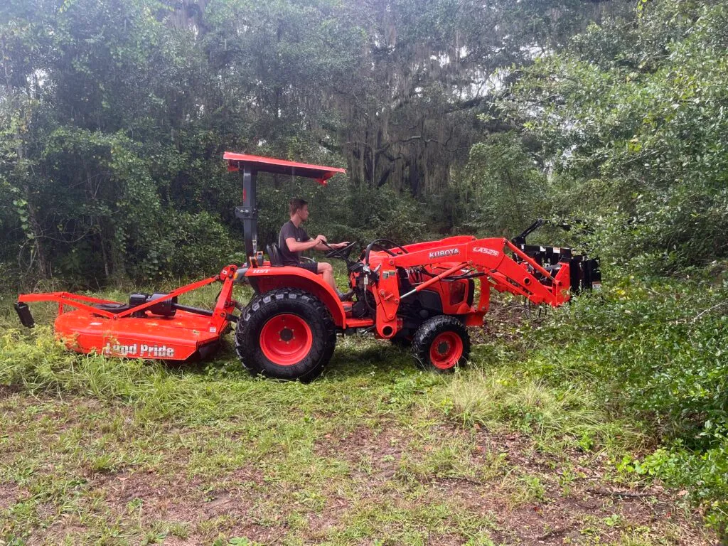 Compact tractor handling brush clearing and site-prep work near a wooded property edge.
